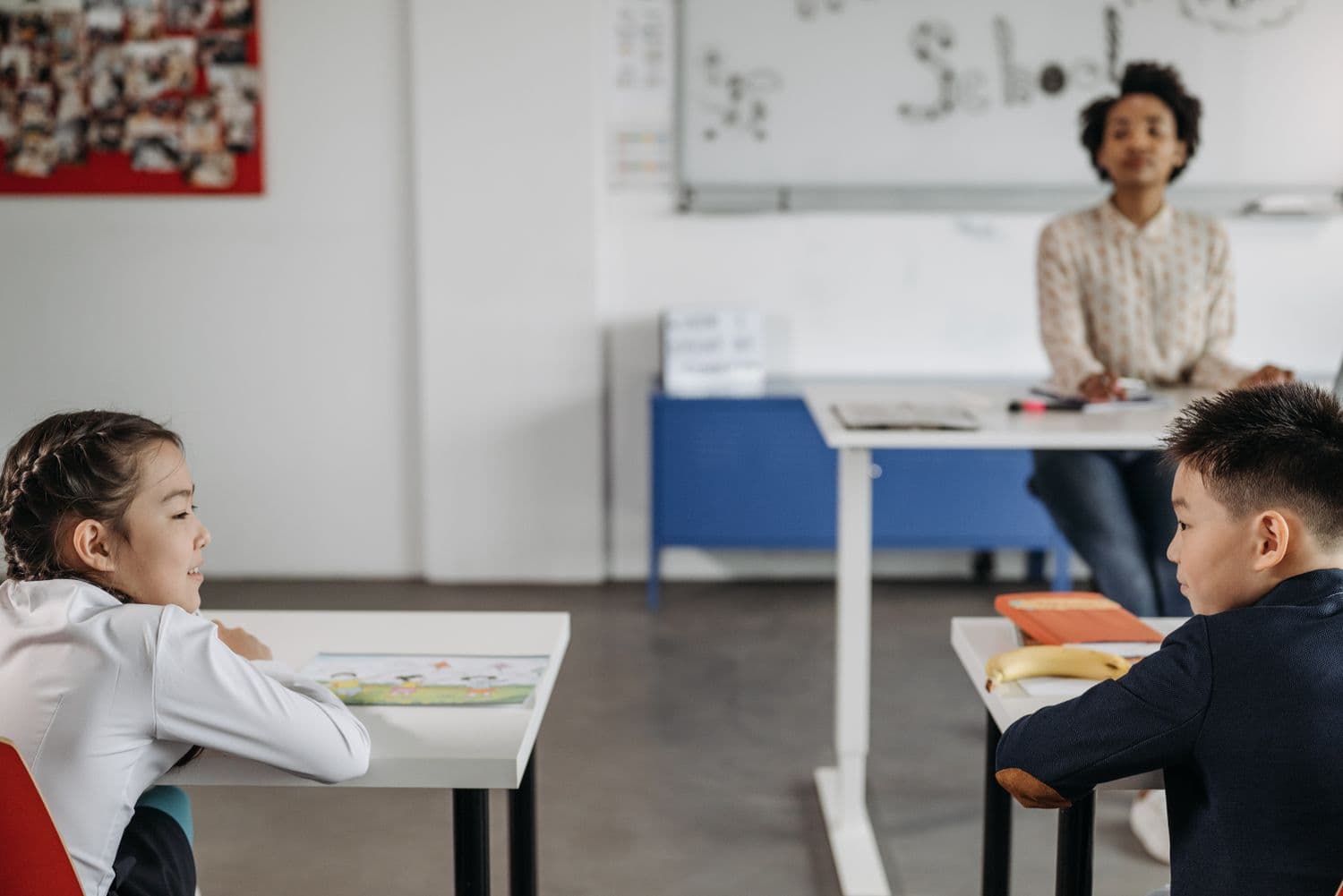 Two young children at their desks in a bright classroom, turning towards their teacher at the front