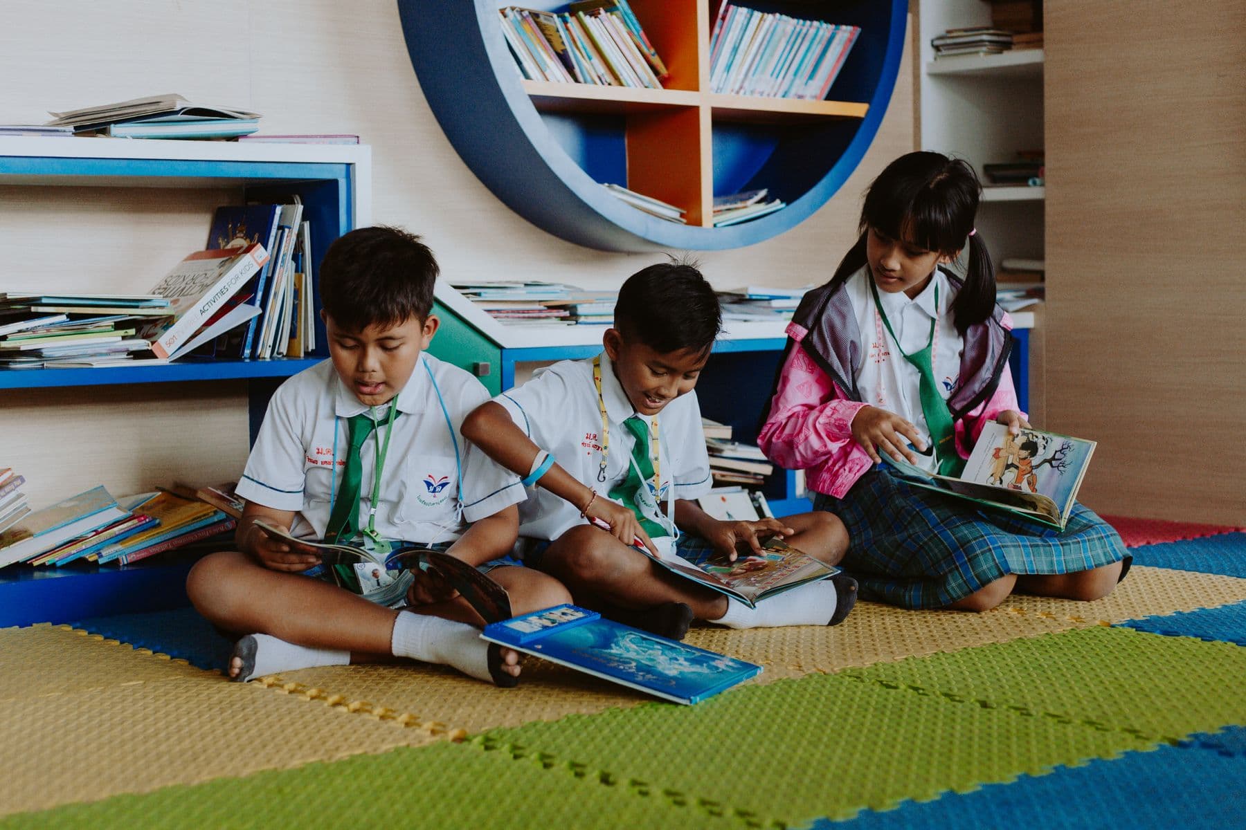 Three primary-age children in school uniforms sitting on a mat reading picture books in a library corner