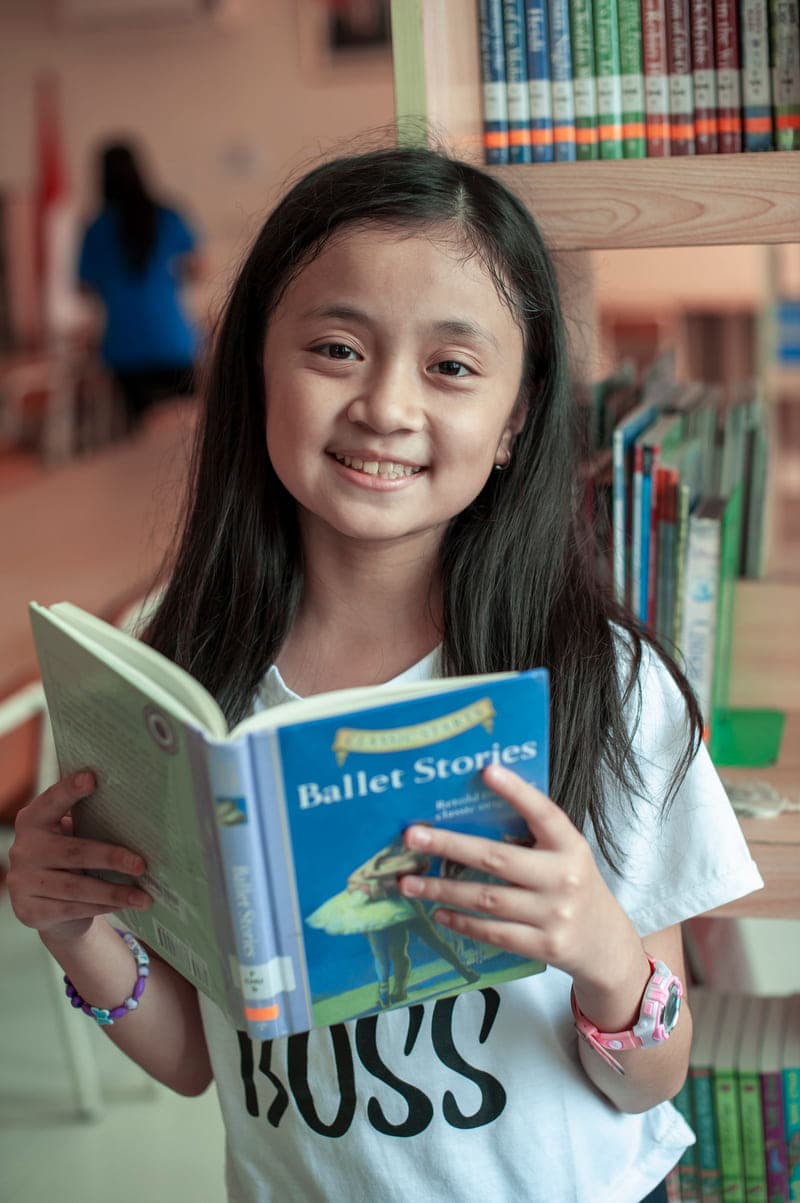 A young student holding an open book and smiling in a school library